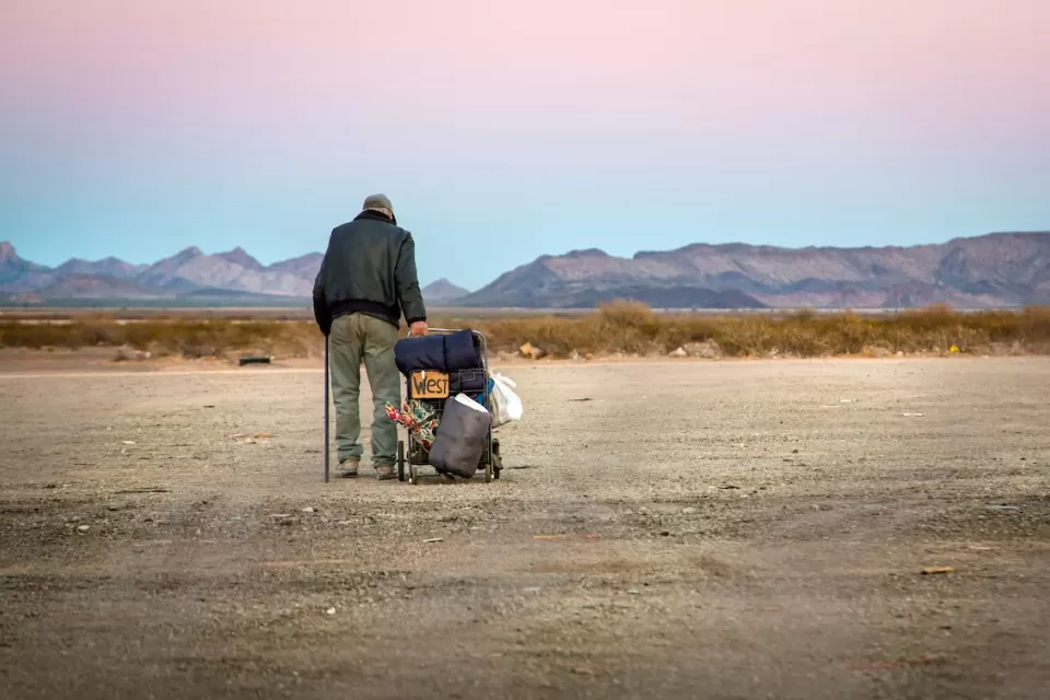 Older unhoused man standing next to cart with belongings and a cardboard sign "West" in a desert landscape.